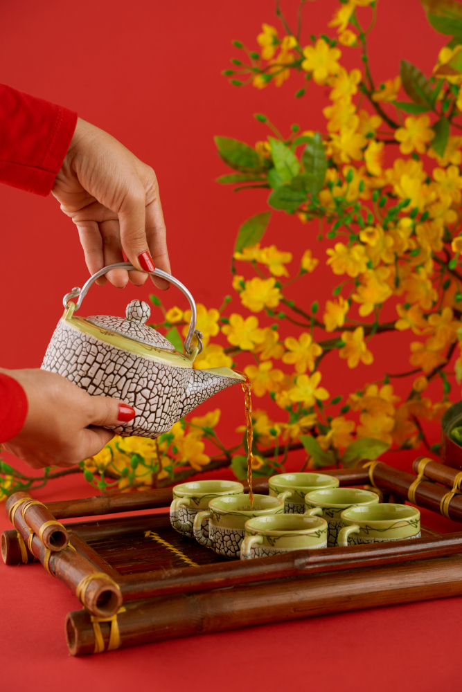 A hand pours tea from a black and white teapot into green cups on a bamboo tray, against a red background with yellow flowers.