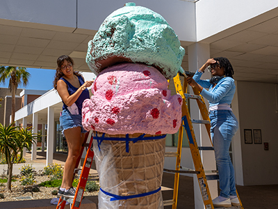 Two women painting an ice cream cone sculpture.