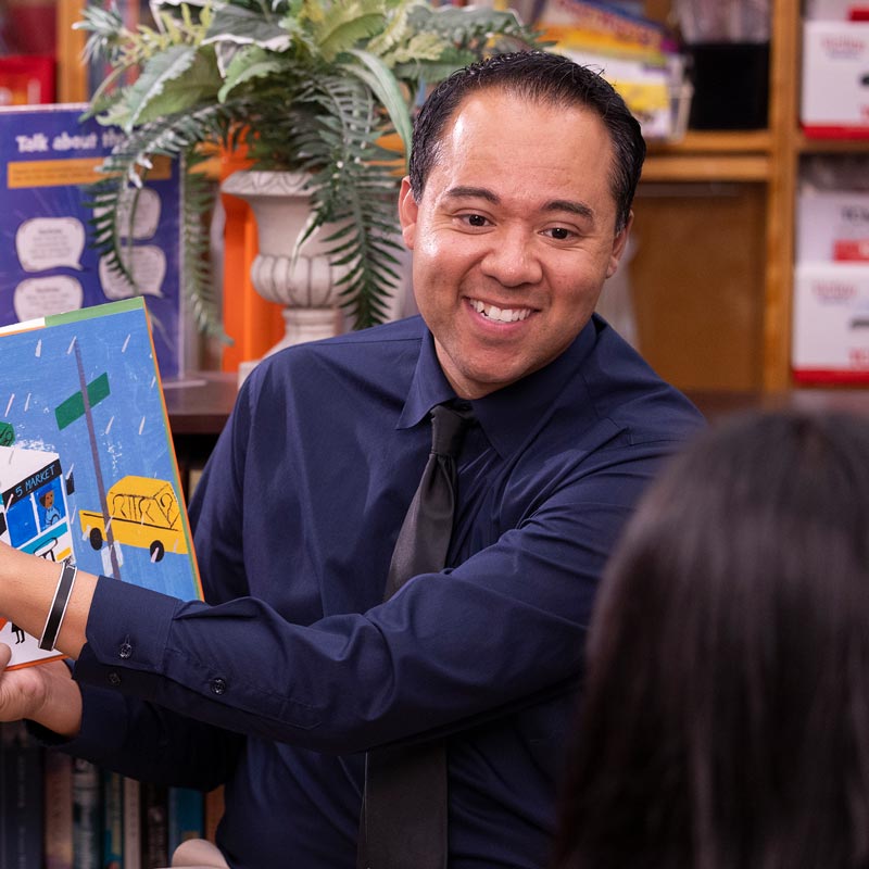 A smiling man in a navy shirt and black tie holds up a colorful children’s book while seated in a classroom or library with a student in the foreground.