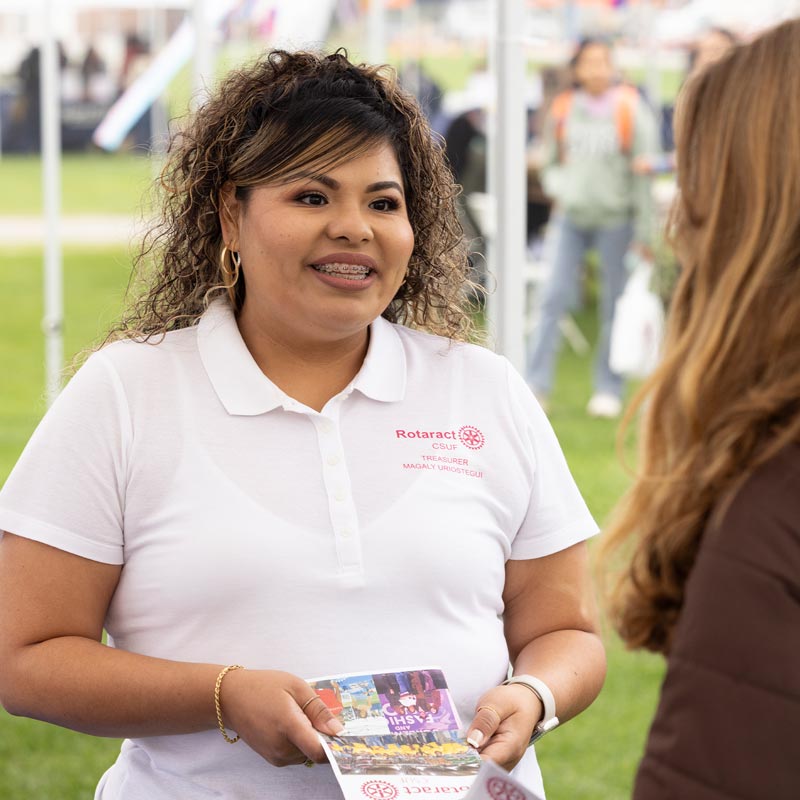 A woman in a white Rotaract polo shirt holds a brochure while speaking with another person at an outdoor event.