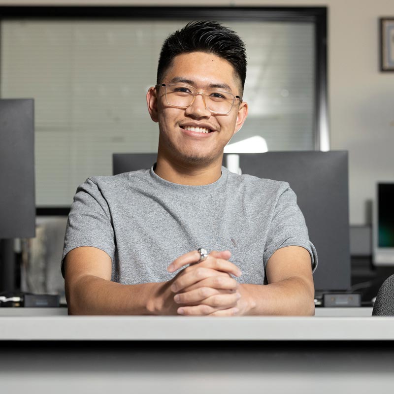 A smiling young man wearing glasses sits at a desk in a computer lab with his hands clasped.