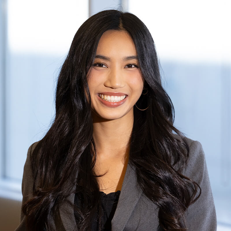 Portrait of a smiling woman with long dark hair wearing a gray blazer in front of softly blurred office windows.