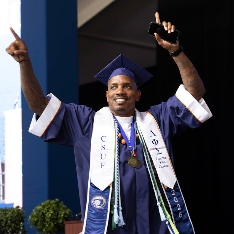 Graduate in blue commencement regalia raises both arms while holding a phone during a graduation ceremony.