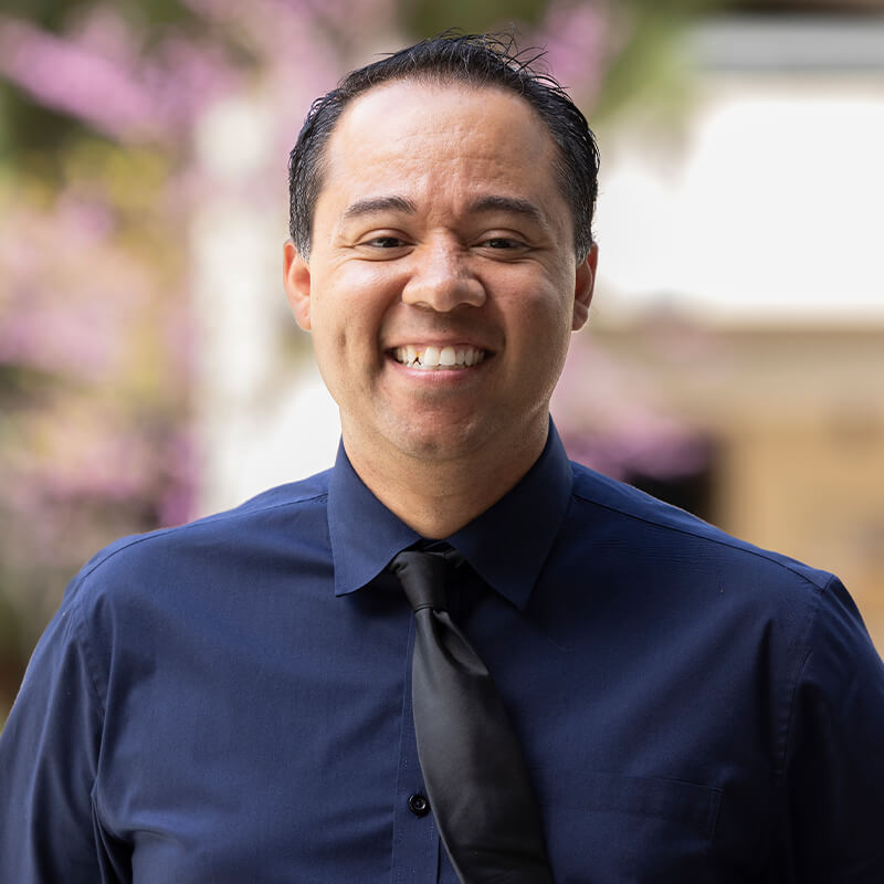 Portrait of a smiling man wearing a navy dress shirt and black tie outdoors with flowering trees blurred in the background.