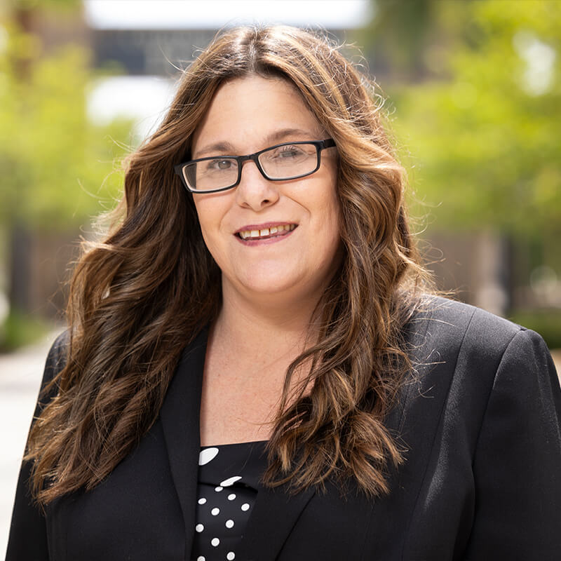 Portrait of a smiling woman with long wavy hair and glasses wearing a black blazer outdoors with a blurred campus background.