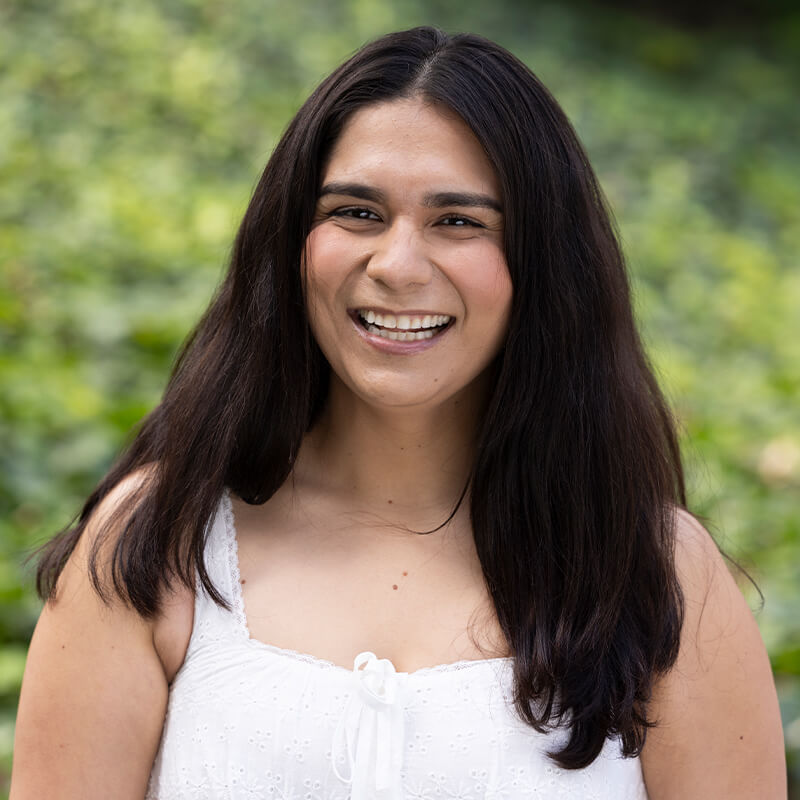 Portrait of a smiling woman with long dark hair wearing a white sleeveless top outdoors with greenery in the background.