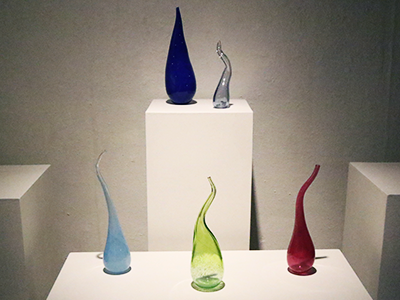 Five blown glass vessels on pedestals.