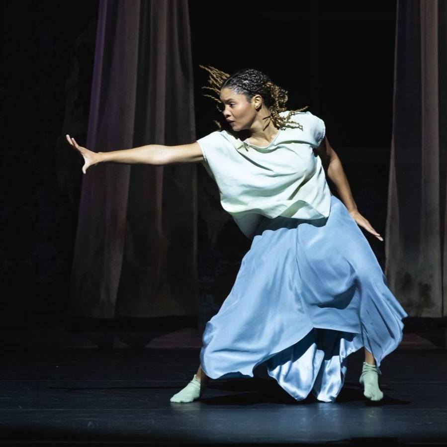 Dancer in a flowing blue skirt reaching towards theleft of the frame. 