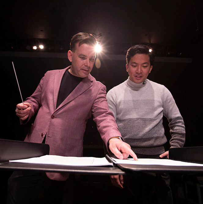 A man with a baton looking at sheet music with another man.