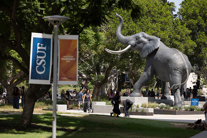 Silhouette of the Titan the Elephant statue with the sun shining through its raised trunk beside a California State University, Fullerton campus building.
