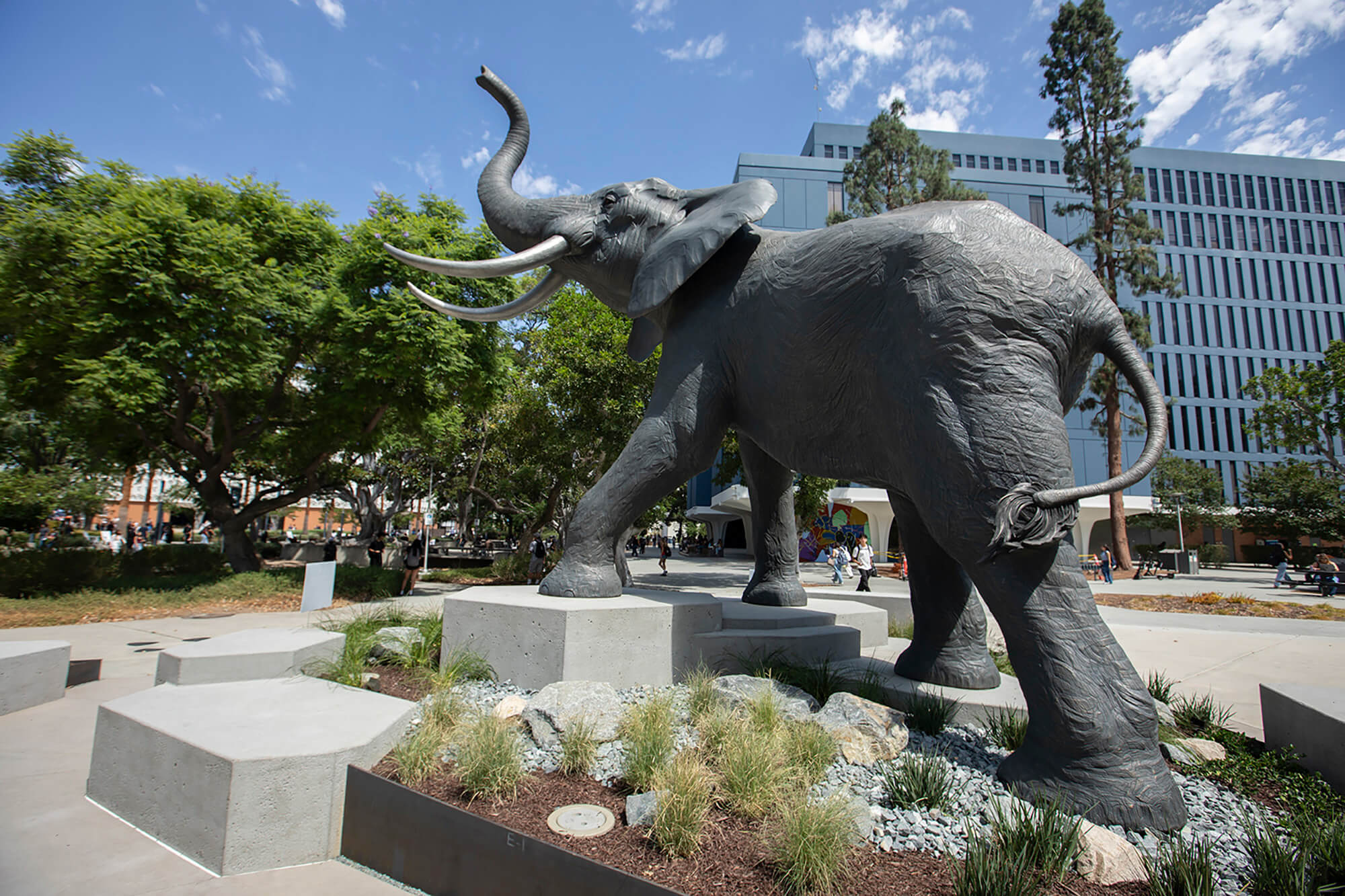 Rear view of the Titan the Elephant statue in a landscaped plaza with campus buildings, trees, and pedestrians at California State University, Fullerton.